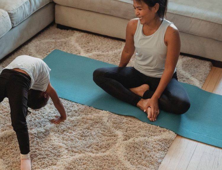 From above of happy mother sitting in lotus pose and looking at daughter bending over backwards at home in daytime