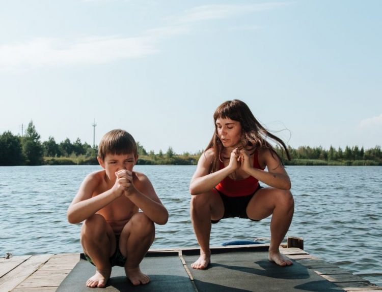 A mother and son exercising on a dock by a serene lake on a sunny day.